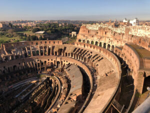 Veduta dell'interno del Colosseo dall'alto, con cielo azzurro e alberi in lontananza.
